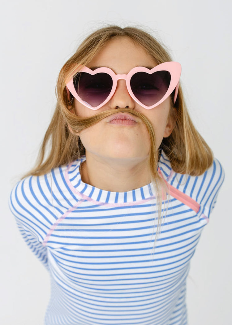 A blonde girl wearing pink heart sunglasses in the striped rashguard from the Cabana Life Girls Palm Beach 3-Piece Long Sleeve Rashguard Set.