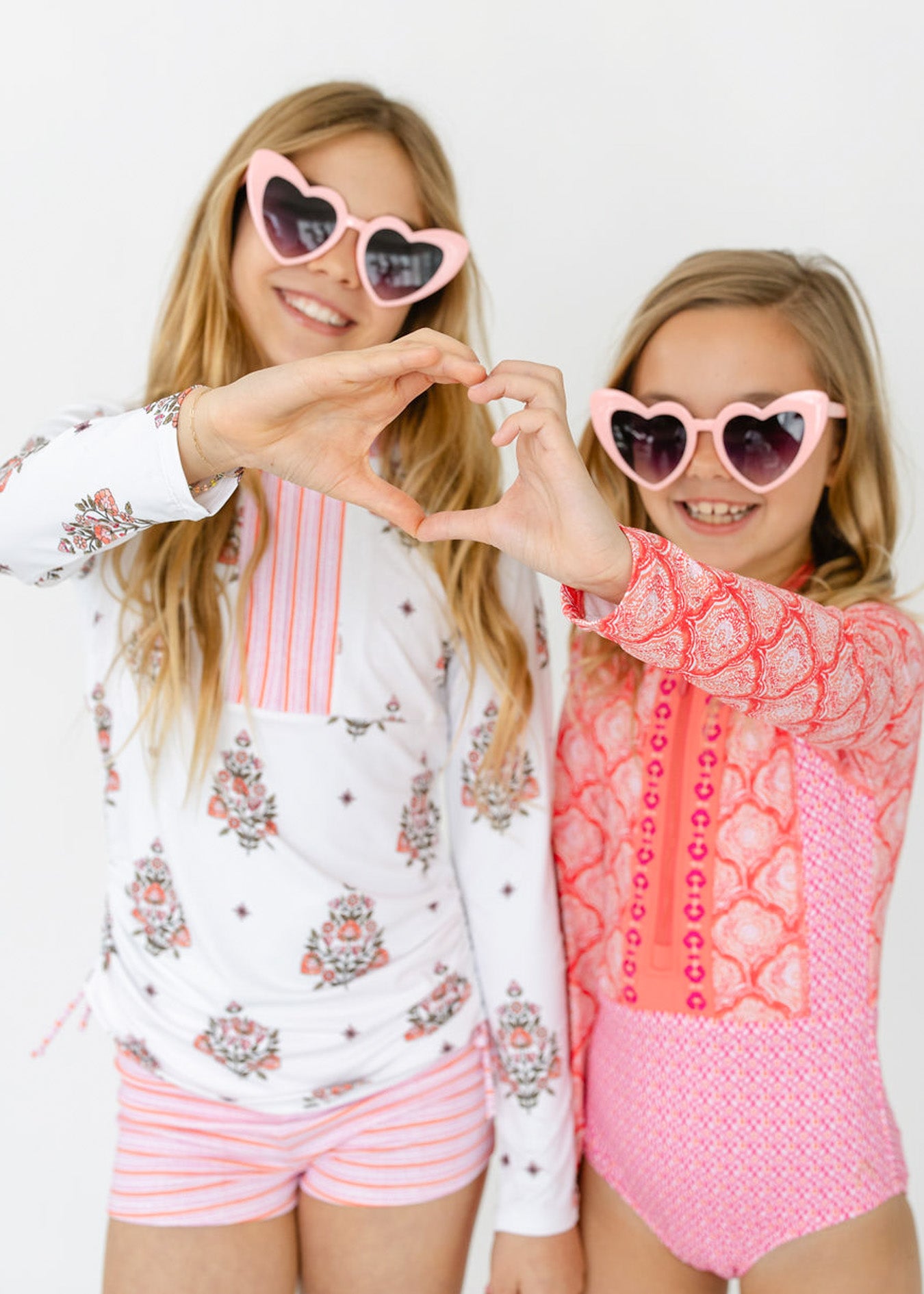 Blonde girls wearing pink heart sunglasses in Cabana Life's sun protective Girls Amelia Island 3-Piece Long Sleeve Rashguard Set and the Girls Amelia Island Long Sleeve One-Piece.