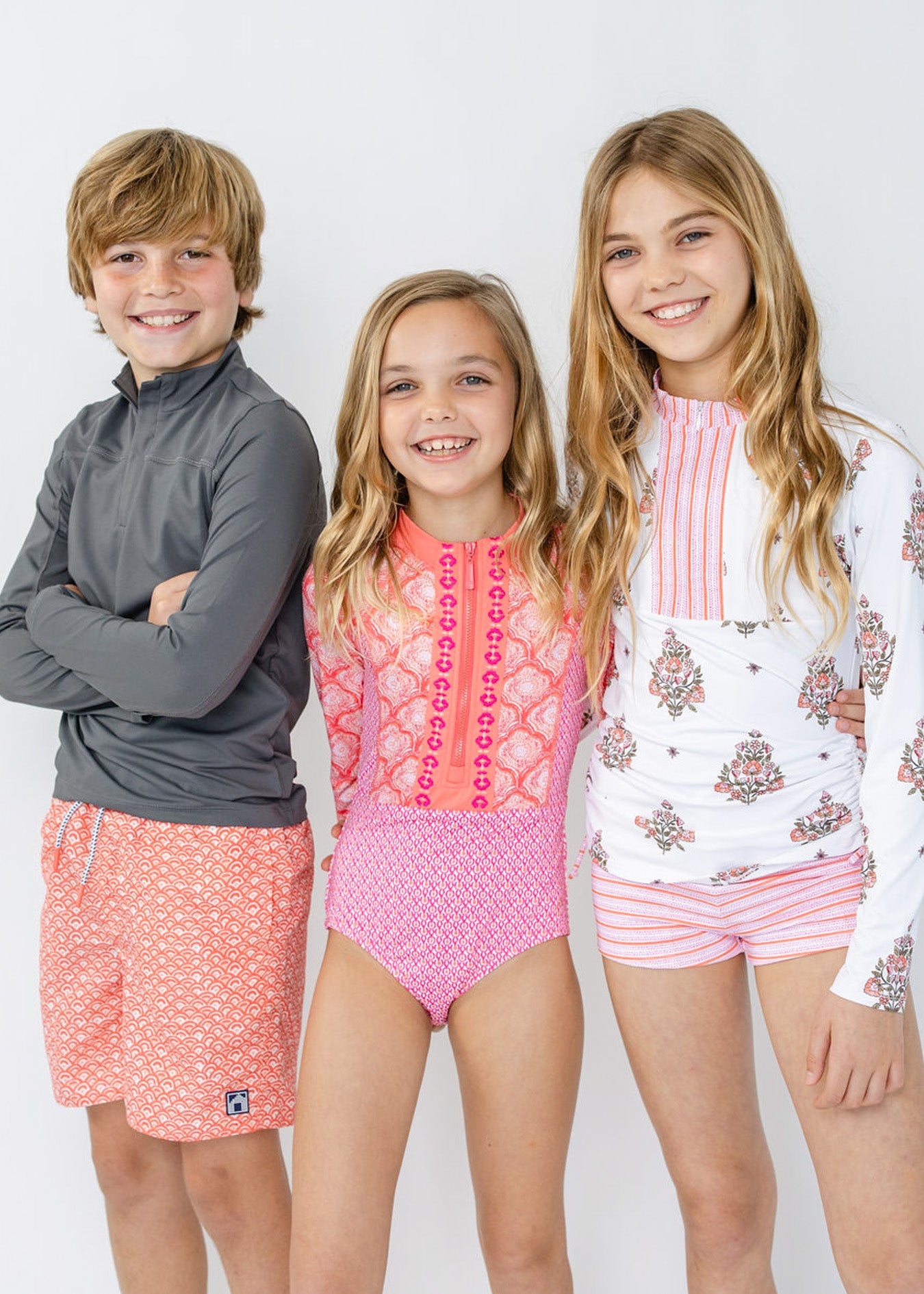 A boy wearing the sun protective Boys Grey Sport Zip Top and Boys Amelia Island Swim Trunk with girls in the Girls Amelia Island 3-Piece Long Sleeve Rashguard Set and Girls Amelia Island Long Sleeve One-Piece.