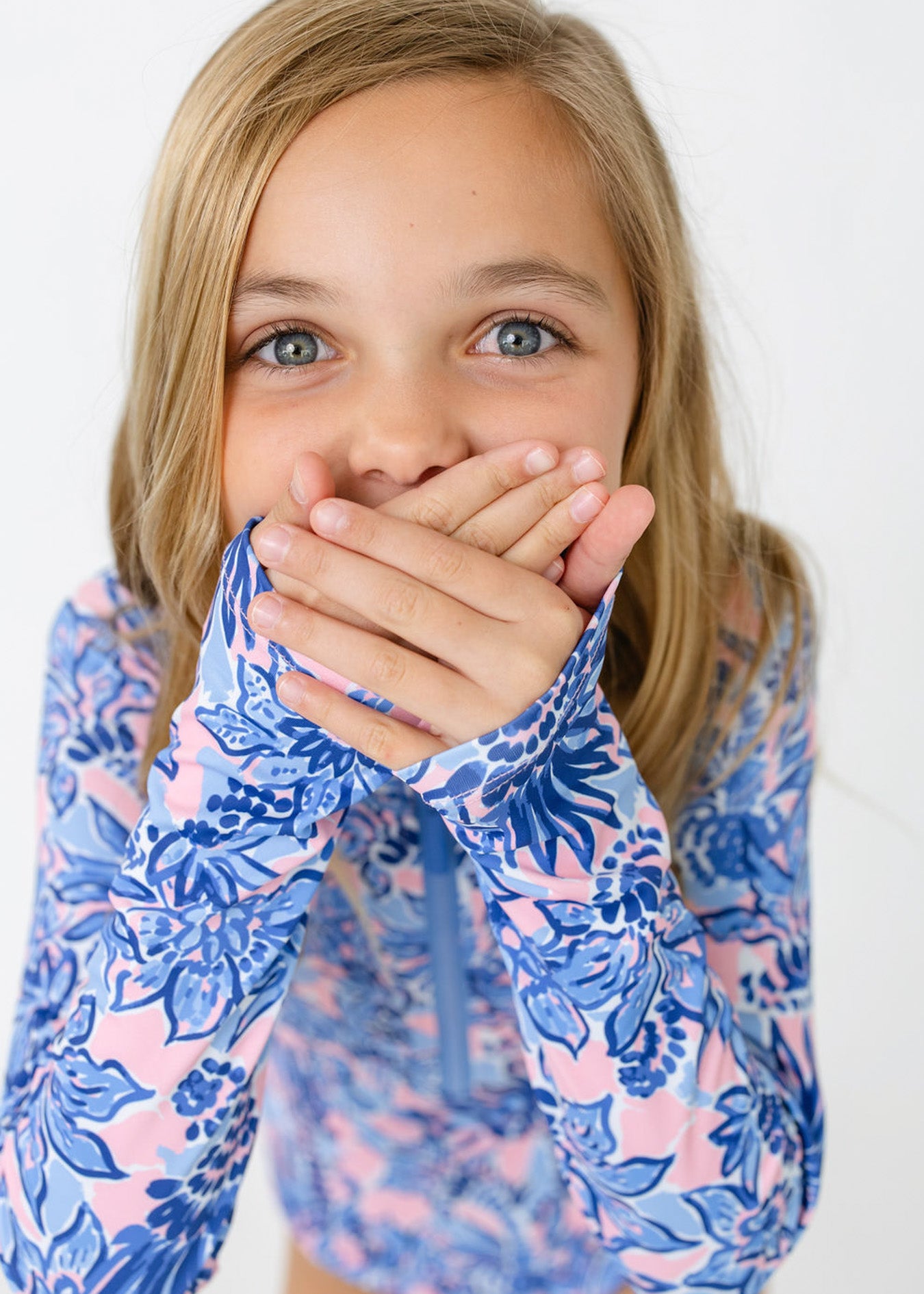 A blonde girl with hands over her mouth wearing the sun protective Girls Palm Beach Long Sleeve One-Piece from Cabana Life.