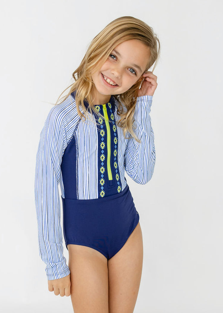 A blonde girl wearing Cabana Life's sun protective Girls Seaside Long Sleeve One-Piece.