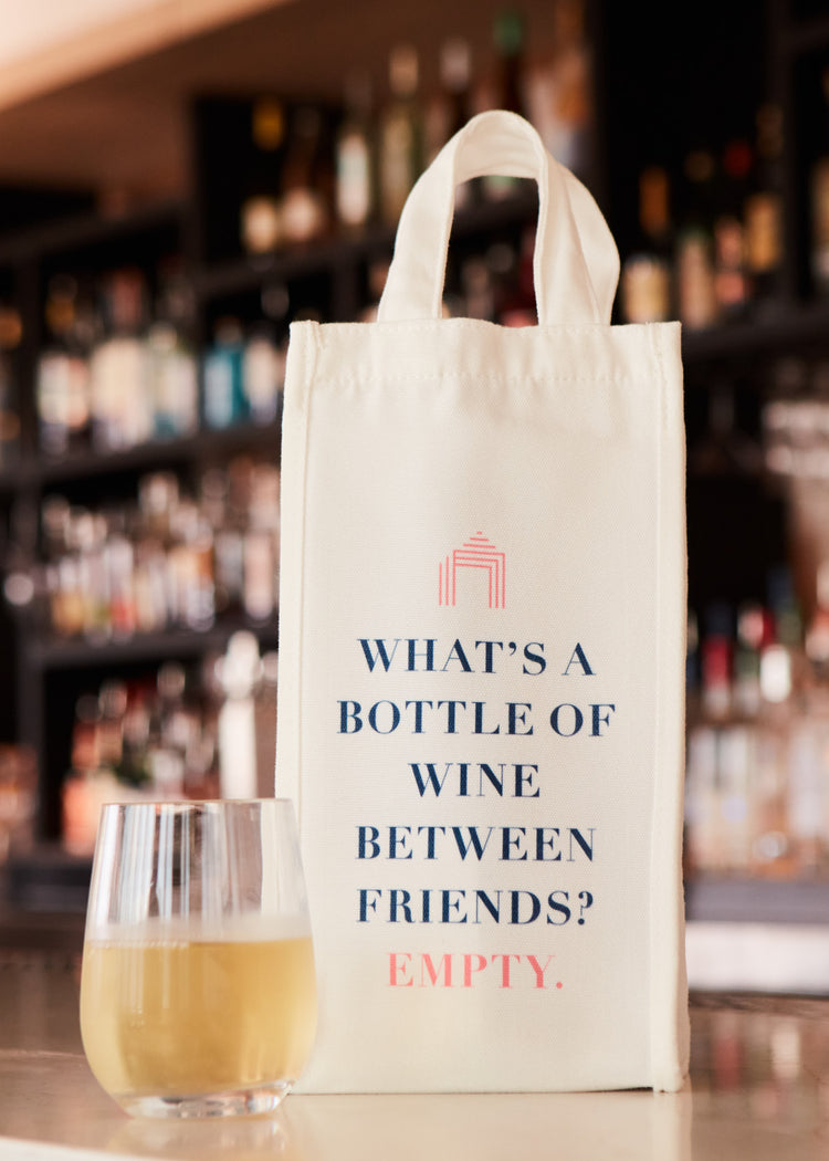 A glass of white wine on a table at a bar with a Cabana Life wine bag that says "What's a bottle of wine between friends? Empty."