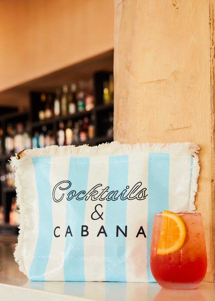 A Cocktails & Cabana Striped Accessory Bag on a bar counter next to an Aperol Spritz glass.