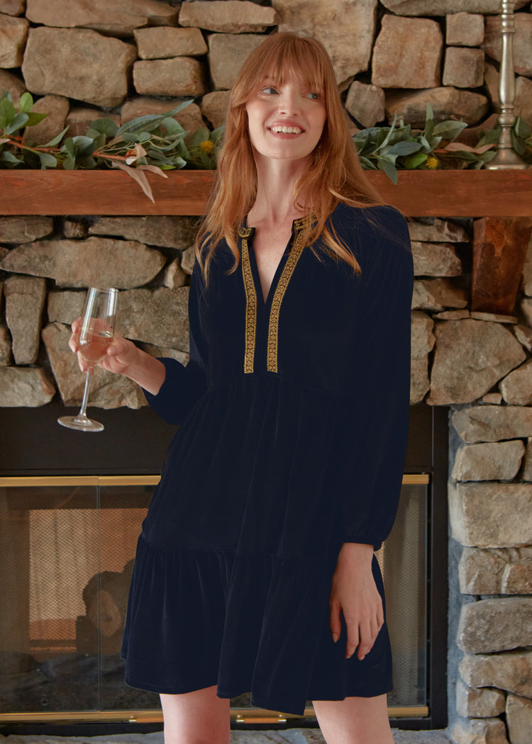 A redhead woman holding a champagne glass in front of a fireplace wearing the Cabana Life sun protective Navy Embroidered Velvet Tiered Dress. 