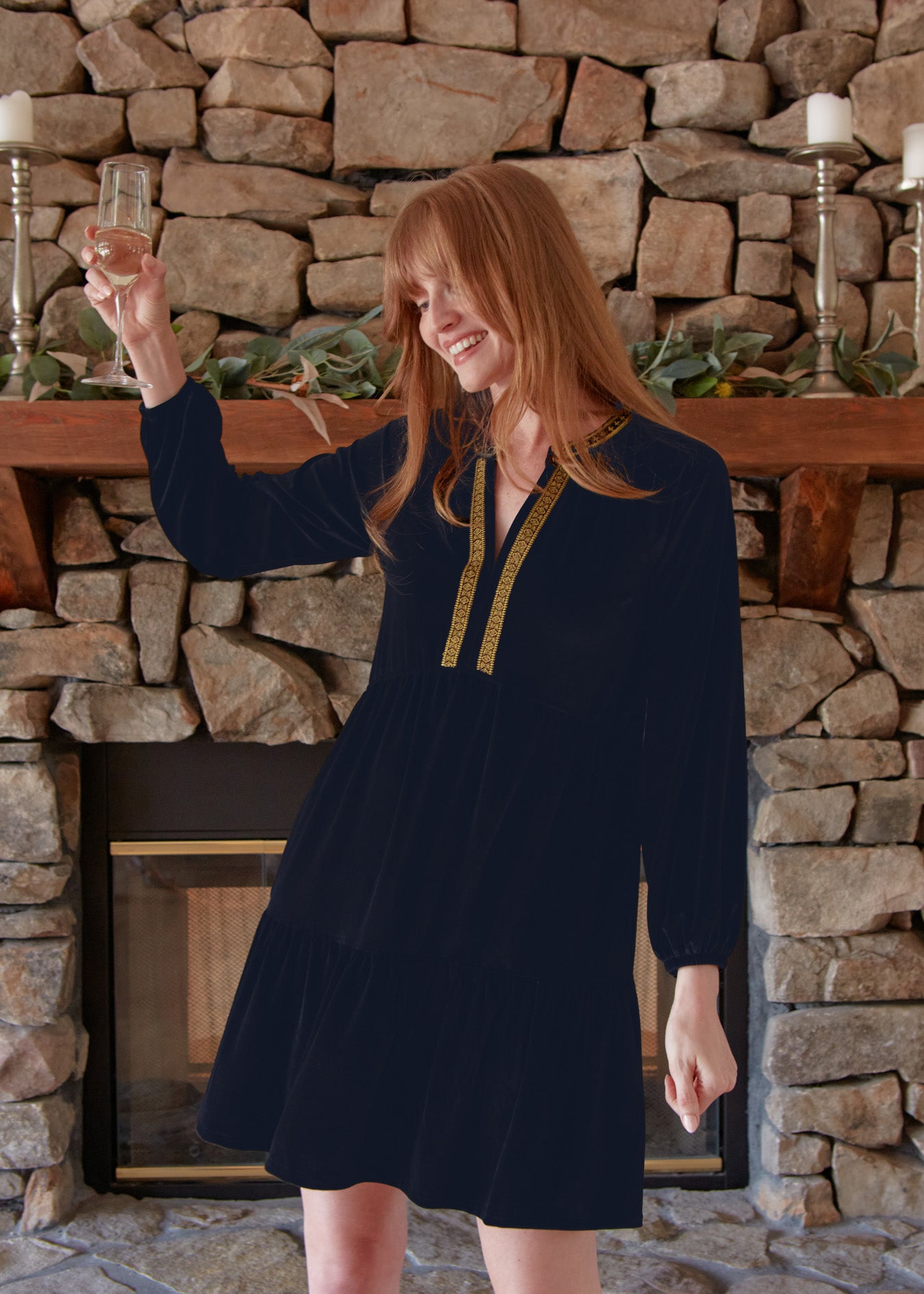 A redhead woman holding a champagne glass up in front of a fireplace wearing the Cabana Life sun protective Navy Embroidered Velvet Tiered Dress. 