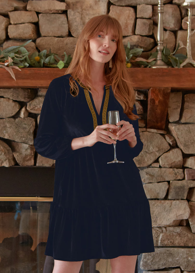 A redhead woman holding a champagne glass in front of her while wearing the Cabana Life sun protective Navy Embroidered Velvet Tiered Dress in front of a fireplace. 