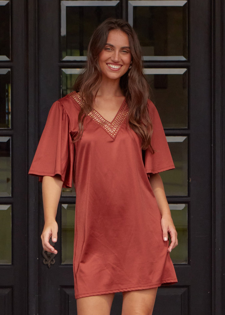 A brunette woman with wavy hair wearing the sun protective Cabana Life Copper Flutter Sleeve Shift Dress walking in front of a black door.