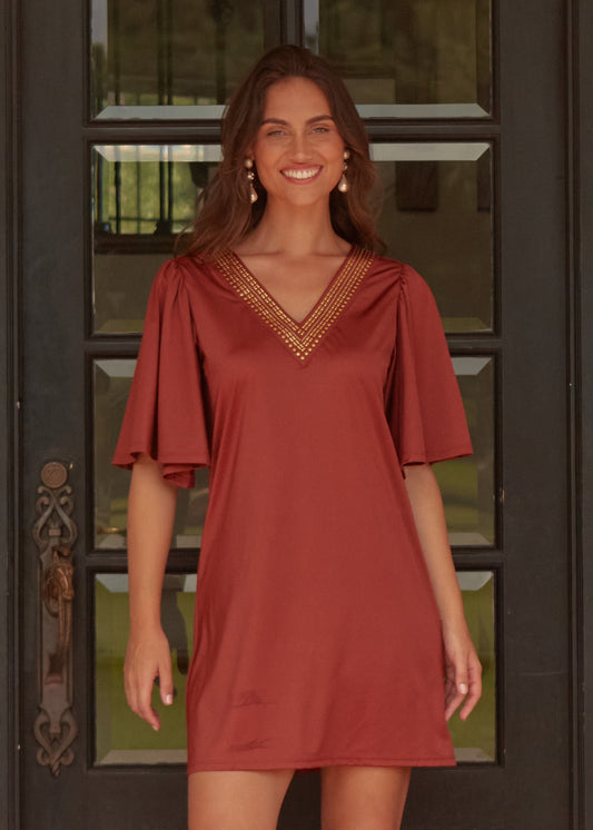 A brunette woman with wavy hair smiling while wearing the sun protective Cabana Life Copper Flutter Sleeve Shift Dress in front of a black door.