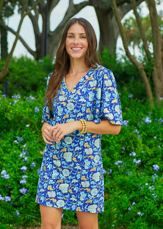 A brunette woman standing in front of a bush with blue flowers wearing the Cabana Life sun protective Navy Floral Metallic Embroidered Flutter Sleeve Shift Dress.