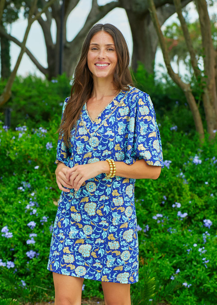 A brunette woman standing in front of a bush with blue flowers wearing the Cabana Life sun protective Navy Floral Metallic Embroidered Flutter Sleeve Shift Dress.