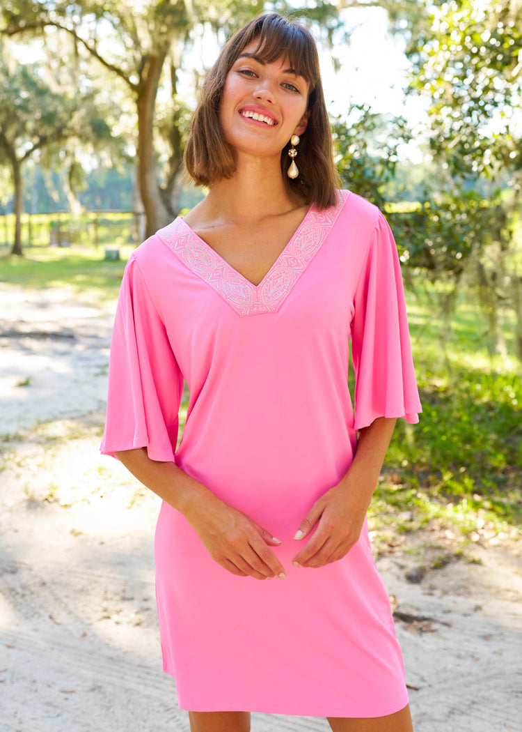 Brunette woman with bobbed hair and bangs standing in front of trees wearing Pink Flutter Sleeve Shift Dress with metallic silver embroidery around v-neck line.