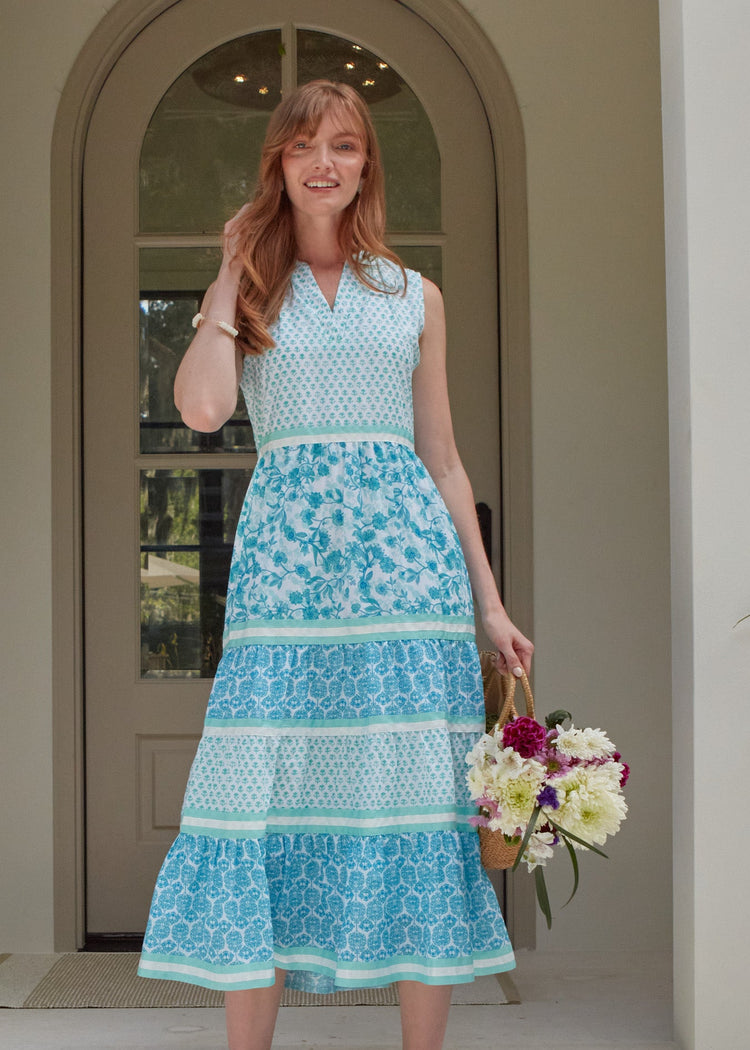 A redhead woman wearing the Cabana Life sun protective Rosemary Beach Sleeveless Tiered Maxi Dress and holding a bag of flowers in front of a house.