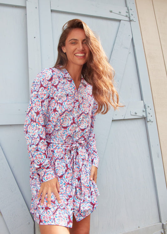 A brunette woman smiling and leaning forward while wearing the sun protective Cabana Life Camden Peony Button Down Shirt Dress in front of a light blue door.