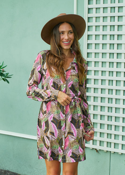 A brunette woman wearing a brown felt halt and tying the waist of Cabana Life's sun protective Myers Button Down Shirt Dress in front of a green building.