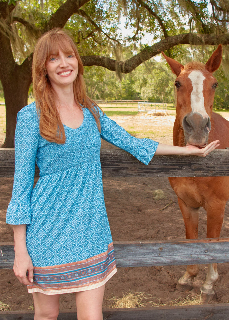 A redhead woman with her arm out feeding a brown horse while wearing the sun protective Cabana Life Camden Leaf Smocked Babydoll Dress.