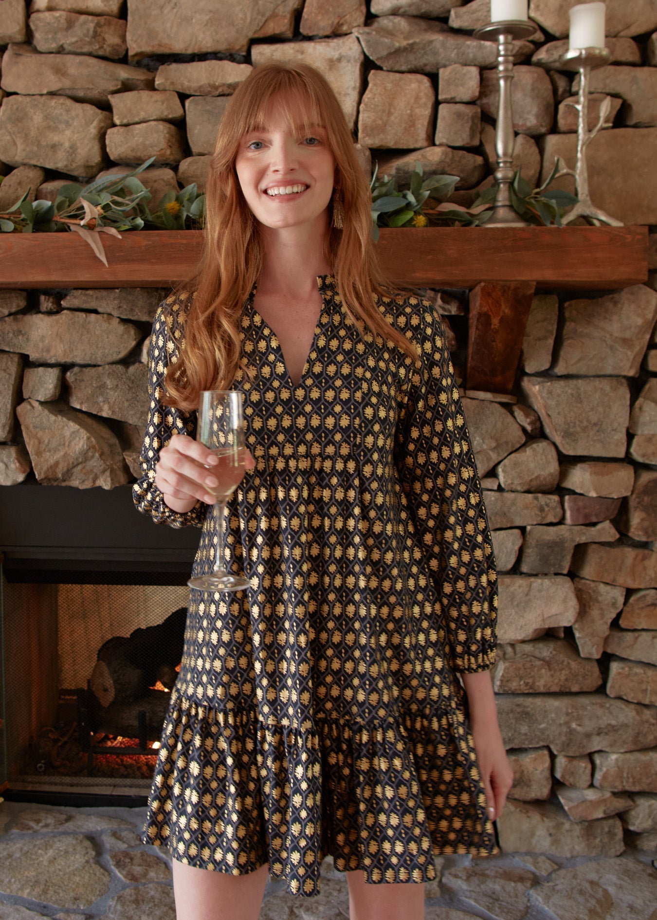 A redhead woman smiling and wearing the sun protective Cabana Life Black Metallic Relaxed Dress in front of a fireplace while holding a glass of champagne.