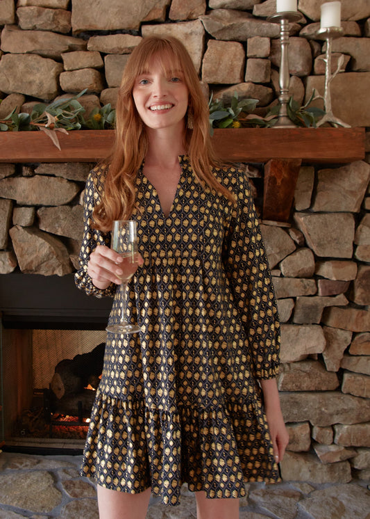 A redhead woman smiling and wearing the sun protective Cabana Life Black Metallic Relaxed Dress in front of a fireplace while holding a glass of champagne.