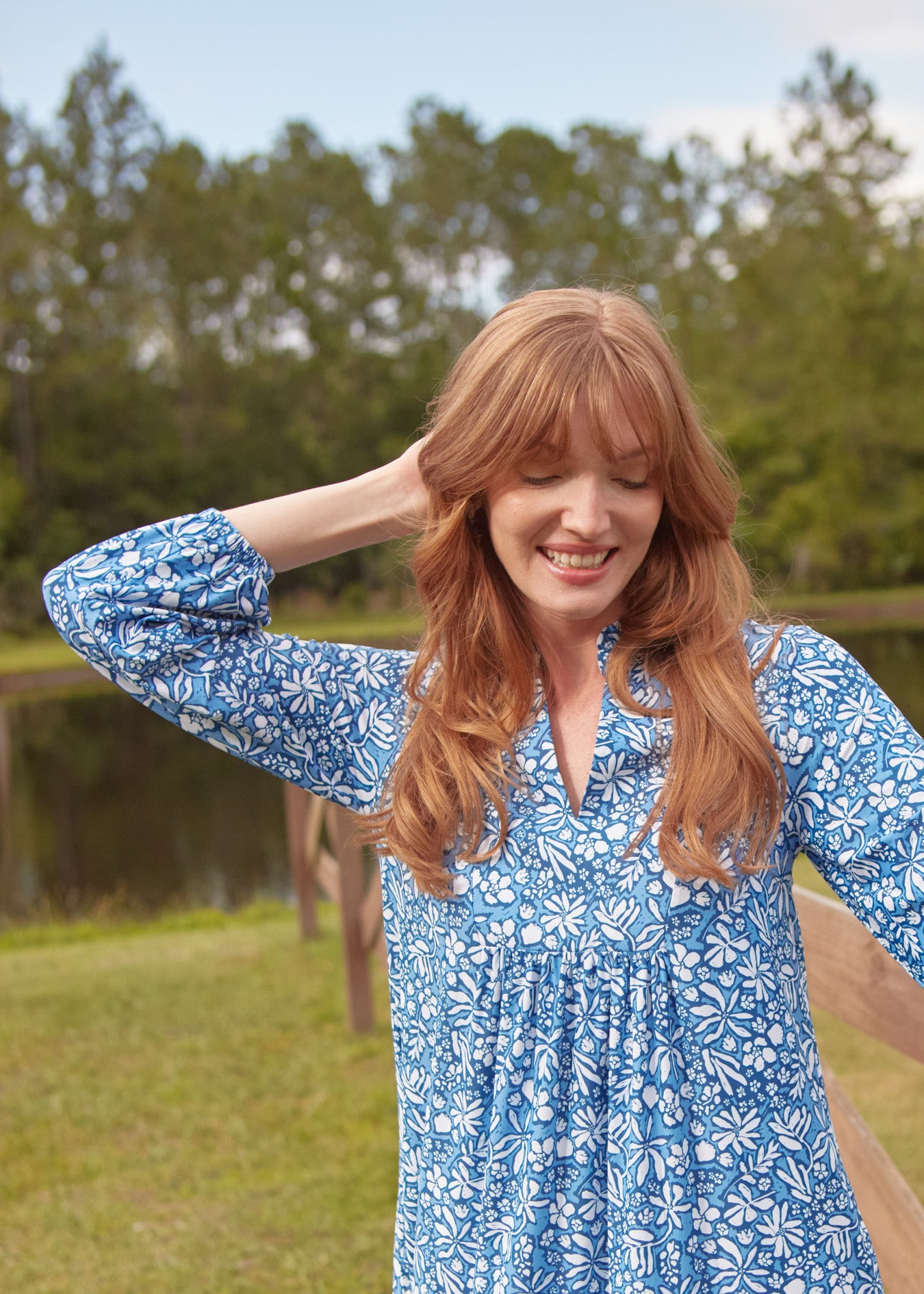 A redhead woman looking down while wearing Cabana Life's sun protective Seaside Floral Relaxed Dress walking in front of a lake & woods.