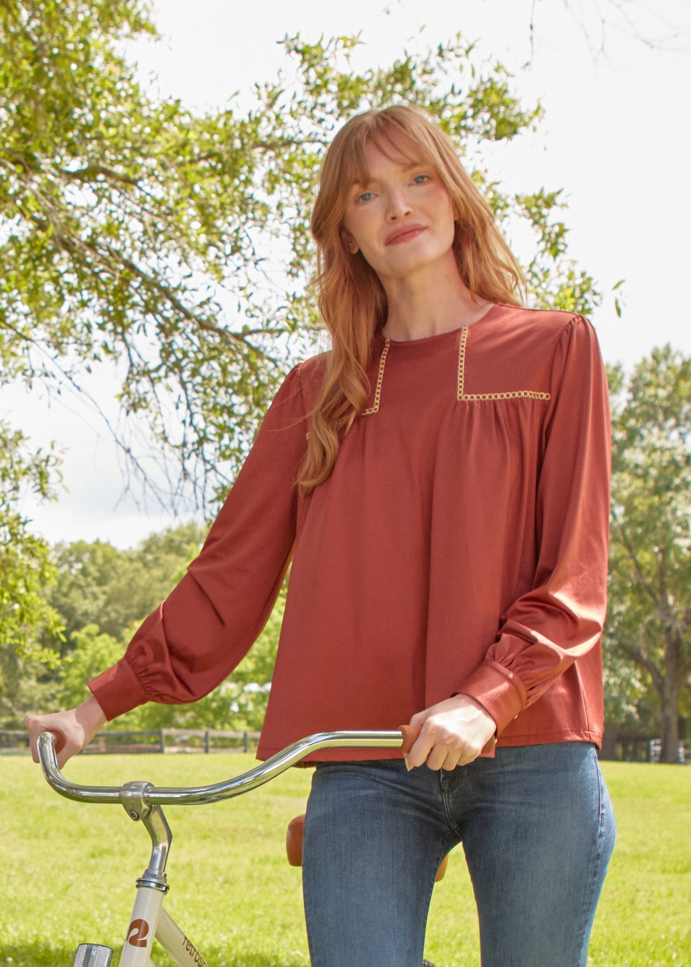 A redhead woman wearing the sun protective Copper Long Sleeve Top from Cabana Life with jeans holding a bike on the grass.