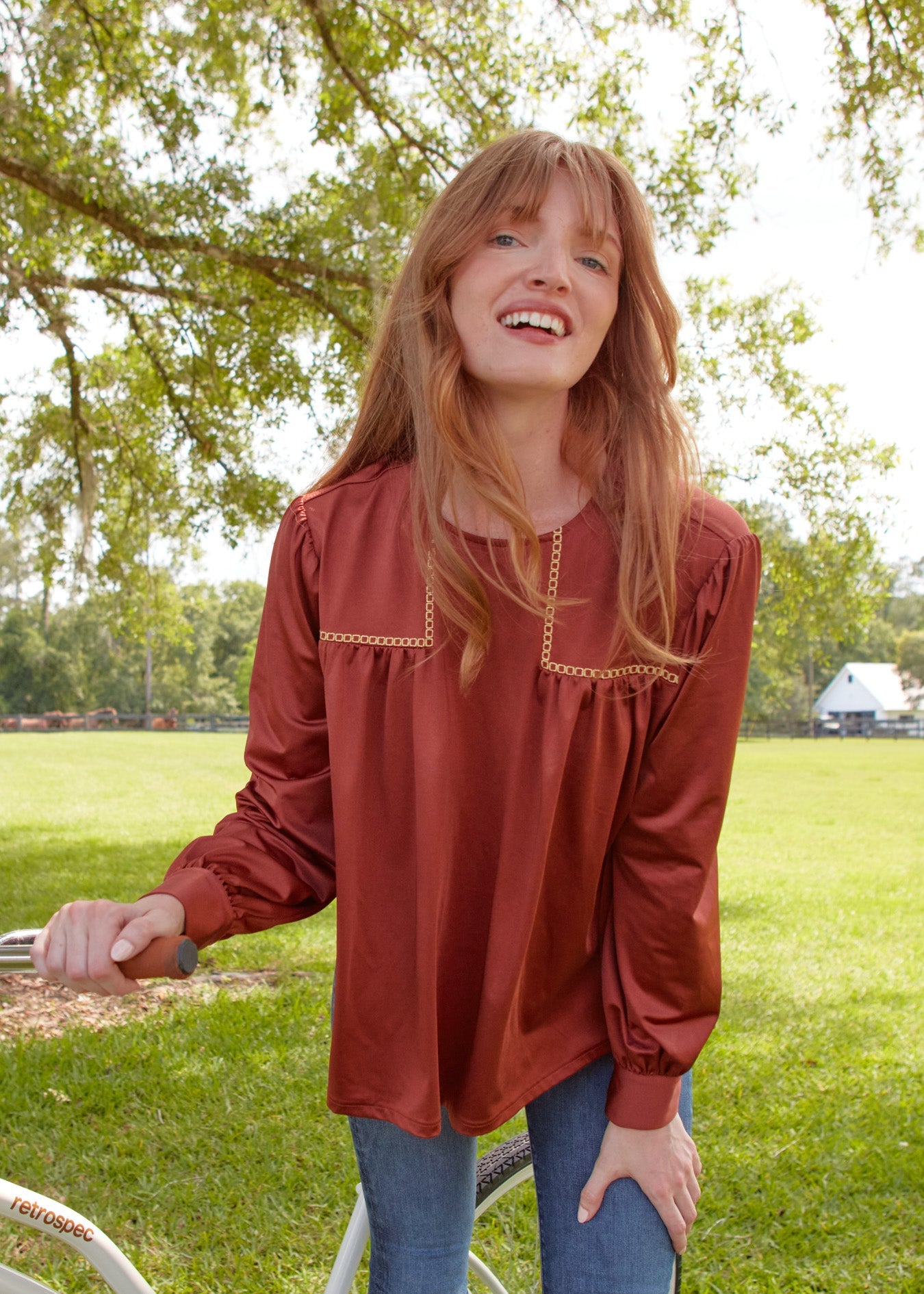 A close up of a readhead woman smiling while wearing the sun protective Copper Long Sleeve Top from Cabana Life with jeans on grass.