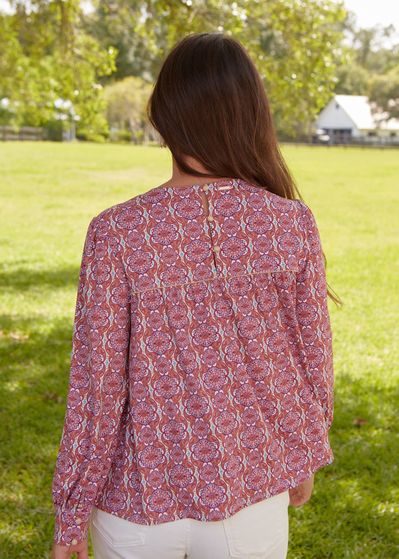 The back of a brunette woman with hair pulled to the front wearing the sun safe Cabana Life Camden Geo Long Sleeve Top with white pants.