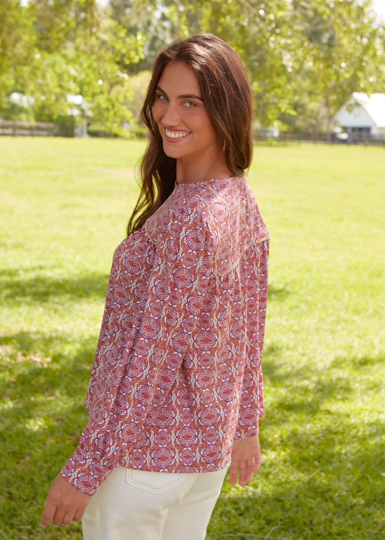 A brunette woman with her head turned smiling and wearing the sun safe Cabana Life Camden Geo Long Sleeve Top with white pants.