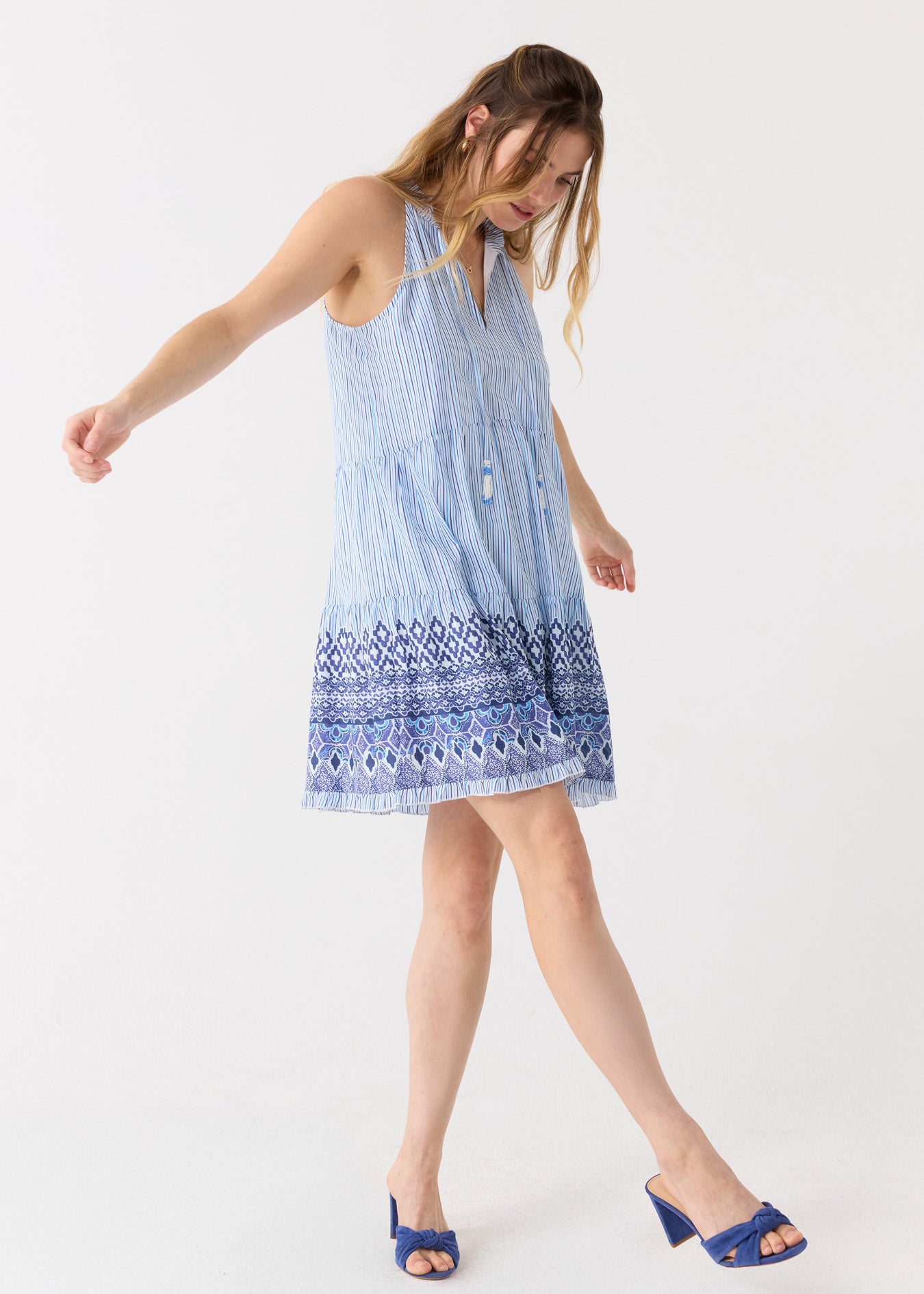 A blonde woman in the Cabana Life sun safe Seaside Sleeveless Tiered Dress  kicking her leg out in blue heels.