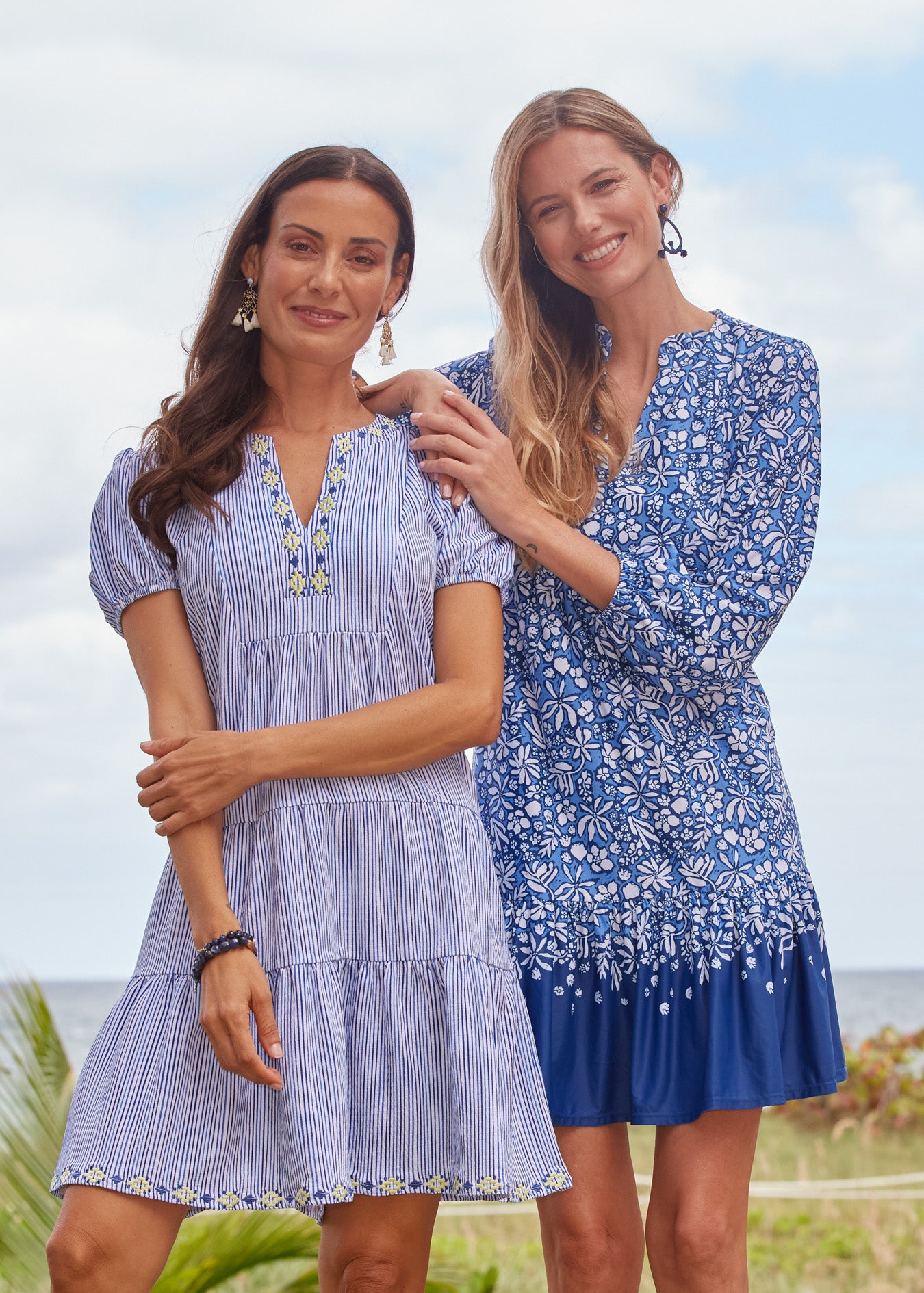 A brunette woman wearing the Cabana Life sun protective Seaside Embroidered Short Sleeve Tiered Dress with a blonde woman in the Seaside Floral Relaxed Dress leaning on her.