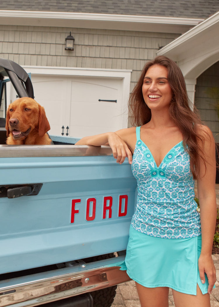 A brunette woman laughing wearing the Cabana Life sun protective Rosemary Beach Embroidered Tankini Top and Aqua Classic Swim Skirt in front of a blue Ford Bronco with a dog.