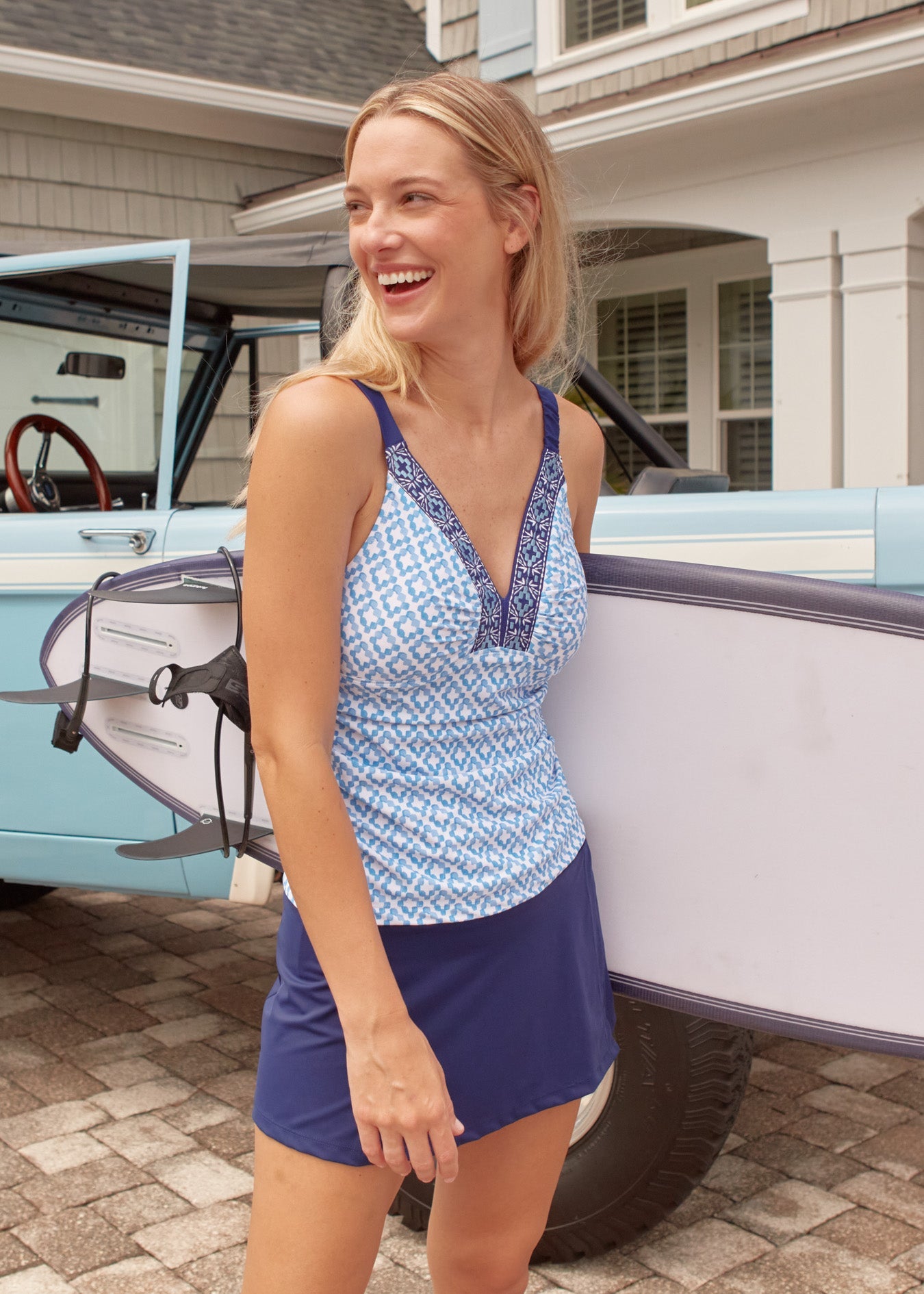 A blonde woman in front of a blue Bronco car holding a surfboard while wearing the Cabana Life sun protective Seaside Embroidered Tankini Top and Navy Classic Swim Skirt.