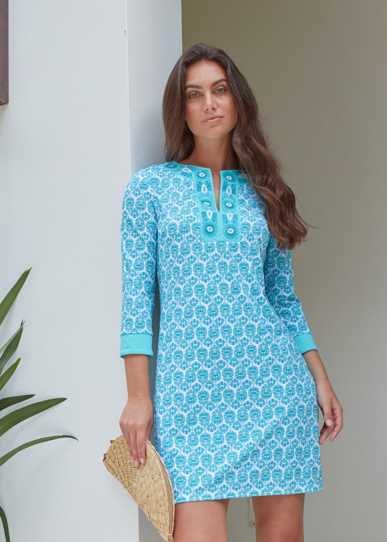 A brunette woman holding a rattan clutch and leaning against a white wall wearing the Cabana Life sun protective Rosemary Beach Embroidered Tunic Dress.