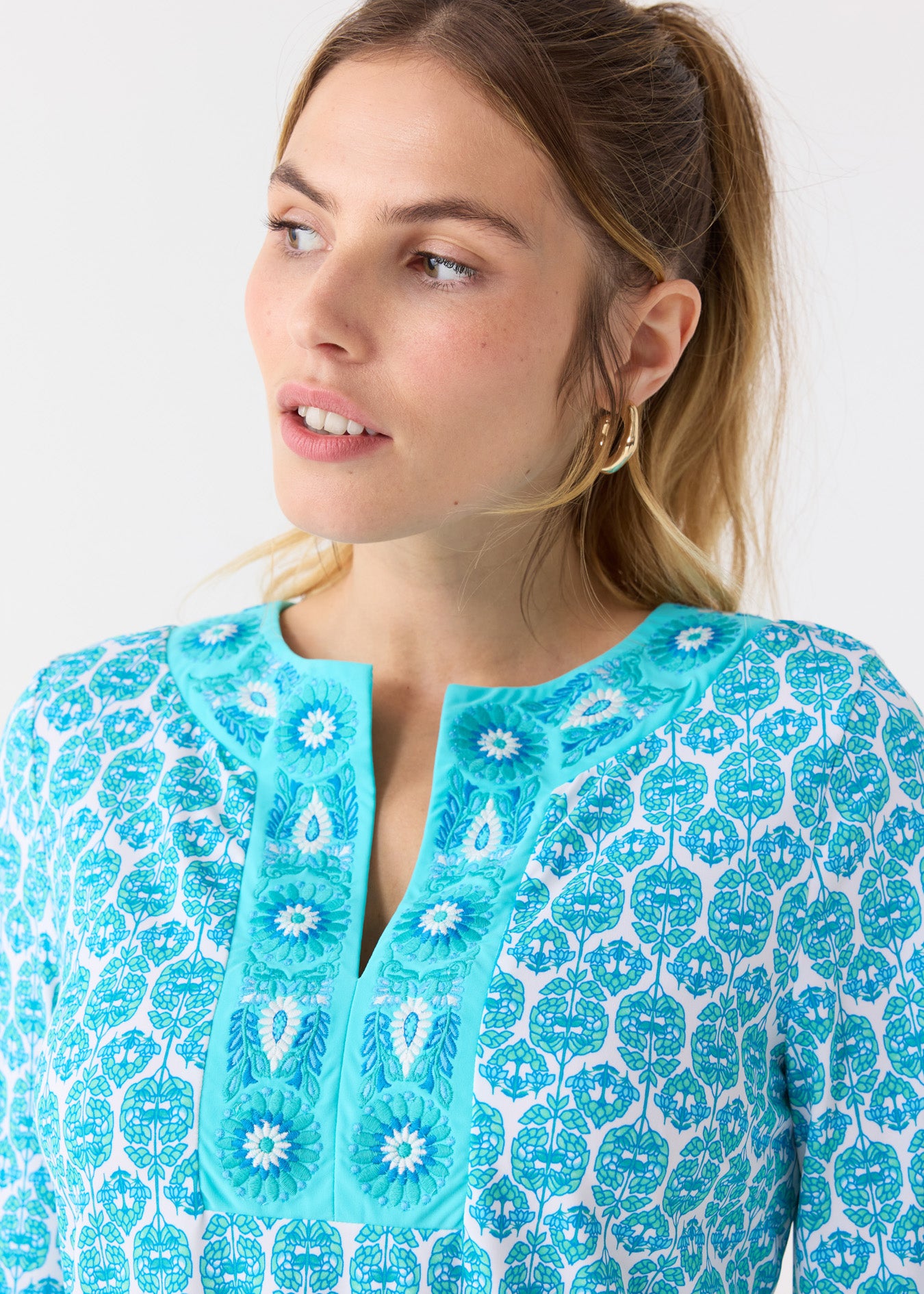 A close up of a woman with her hair up in a ponytail wearing the Cabana Life sun protective Rosemary Beach Embroidered Tunic Dress with gold hoops.