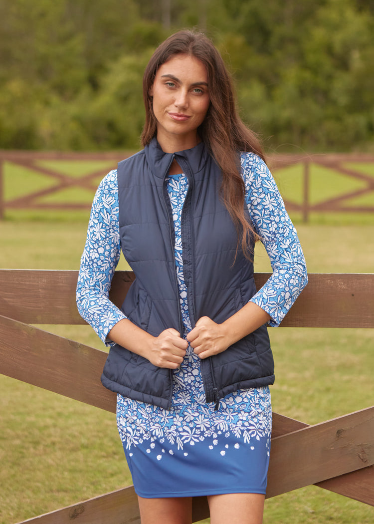 A brunette woman in front of a brown fence and grass wearing Cabana Life's sun protective Seaside Floral Cabana Shift Dress and a navy vest on top.