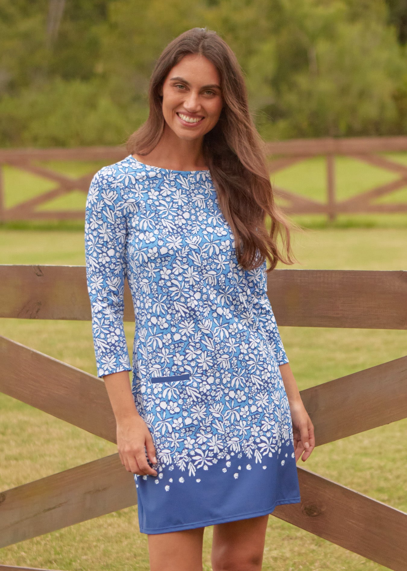 A brunette woman in front of a brown fence and grass wearing Cabana Life's sun protective Seaside Floral Cabana Shift Dress.