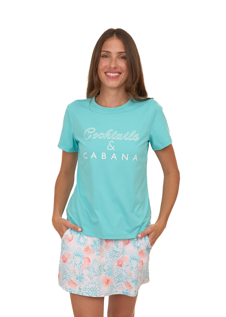 A brunette woman wearing Cabana Life's sun protective Cocktails & Cabana Short Sleeve Top and hands in pockets of the Spritz Skort on a white background.