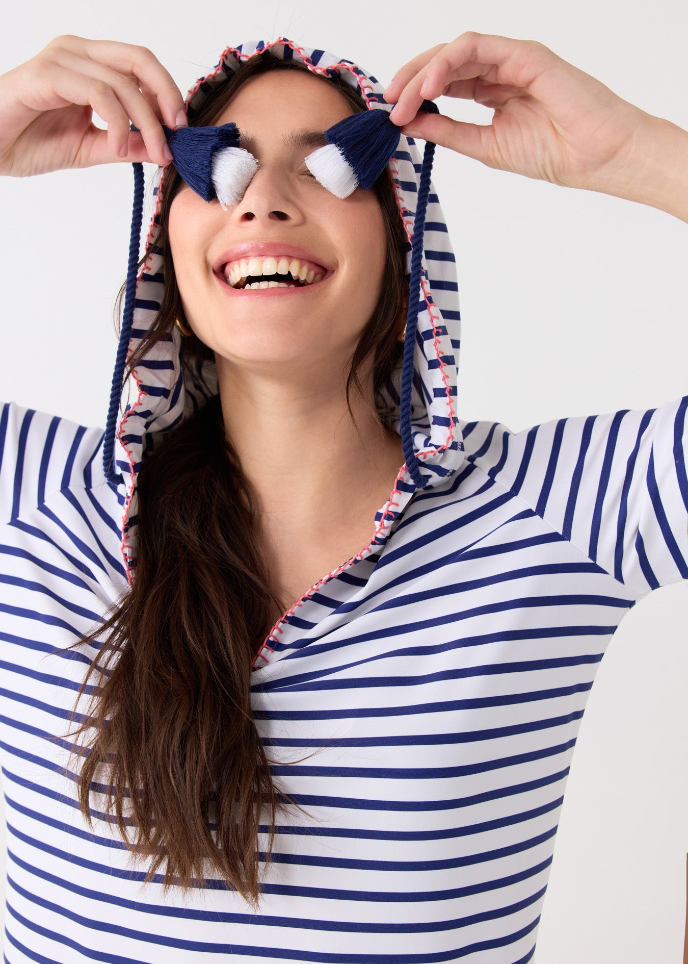 A brunette woman wearing the hood up on the Cabana Life sun protective Navy Stripe Hooded Cover Up and holding the navy and white tassels in front of her eyes.