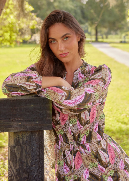 A brunette woman leaning on a fence wearing the sun protective Cabana Life palm print Myers Button Down Maxi Dress.