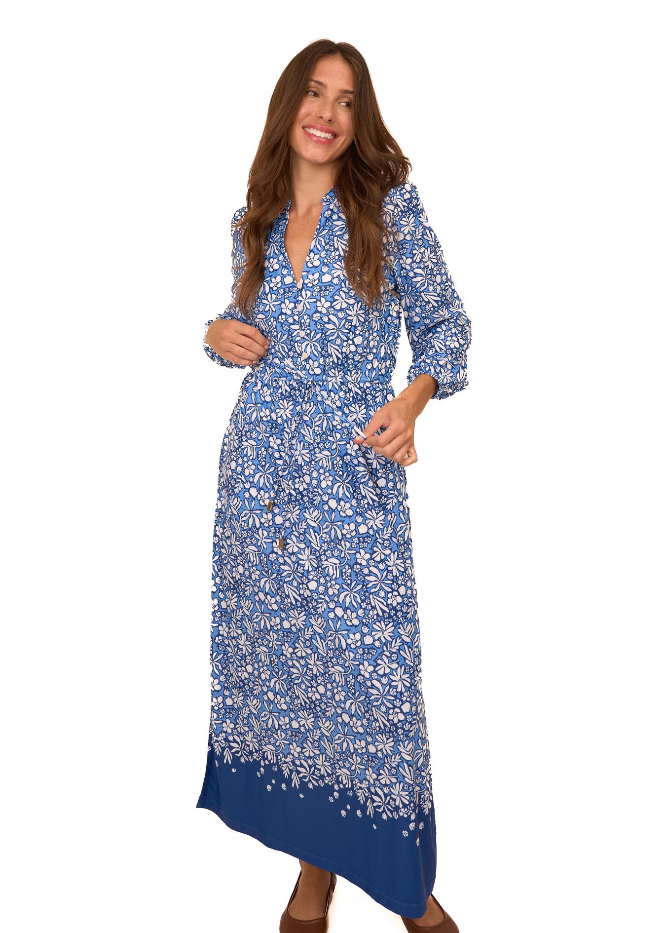 A brunette woman wearing Cabana Life's sun protective Seaside Floral Button Down Maxi Dress and holding the tie waist with brown ballet flats on a white background.