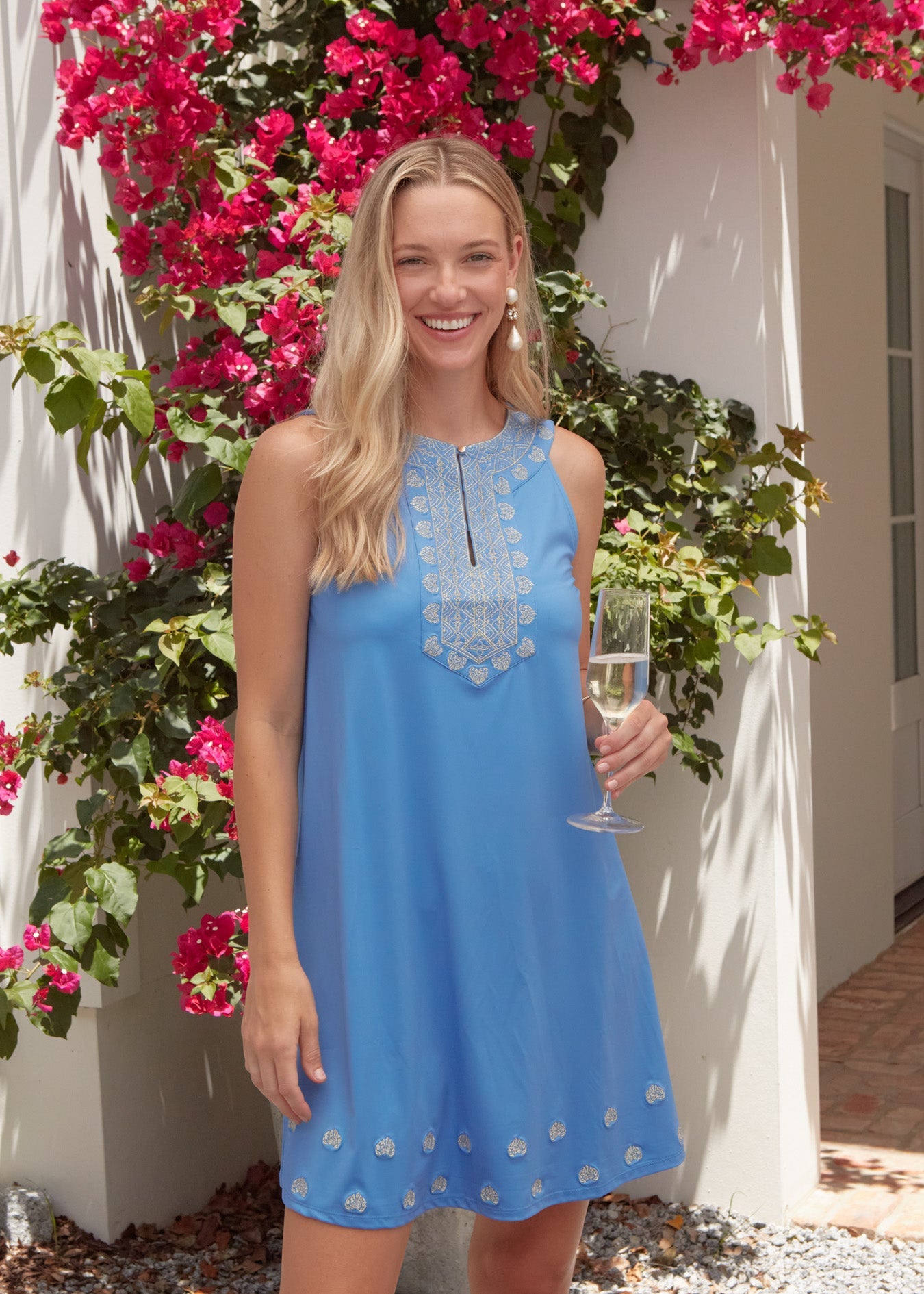 A blonde woman holding a glass of champagne wearing the Cabana Life sun protective Periwinkle Metallic Sleeveless Shift Dress in front of a pink flowers on a wall.