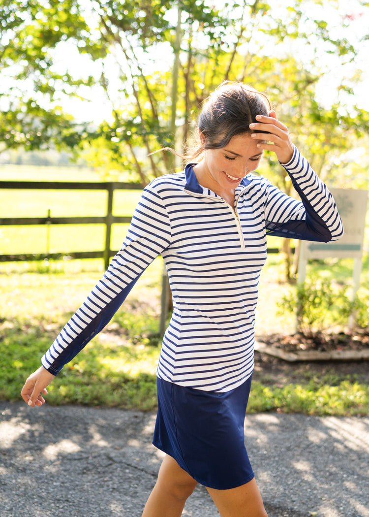 A brunette woman walking while wearing the sun protective Cabana Life Navy Stripe 1/4 Zip Sport Top with Navy 17" Skort in front of greenery.