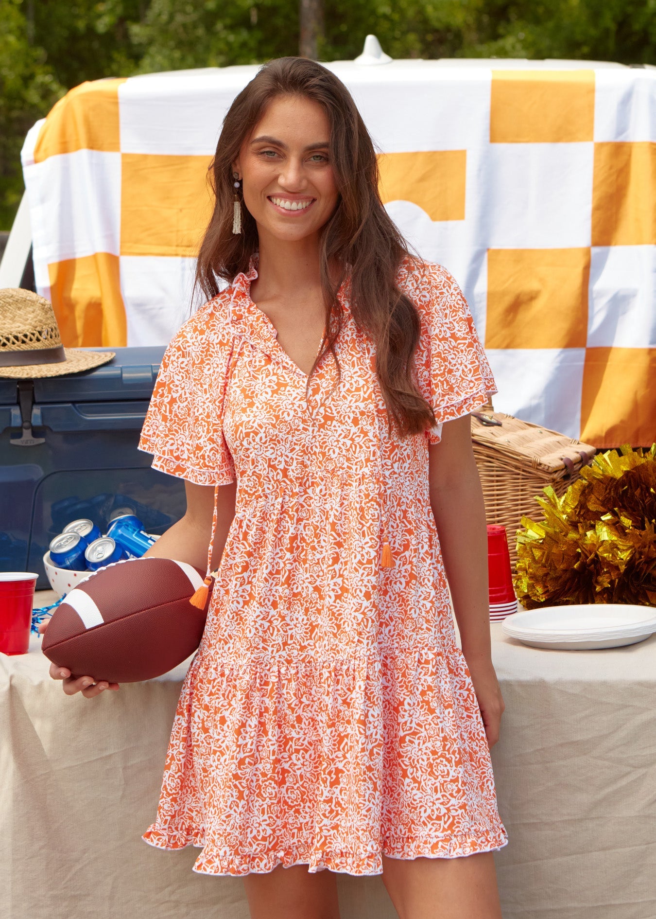 A brunette woman smiling and holding a football in front of a Game Day tailgate truck setup in the sun protective Orange Short Sleeve Tiered Dress from Cabana Life.