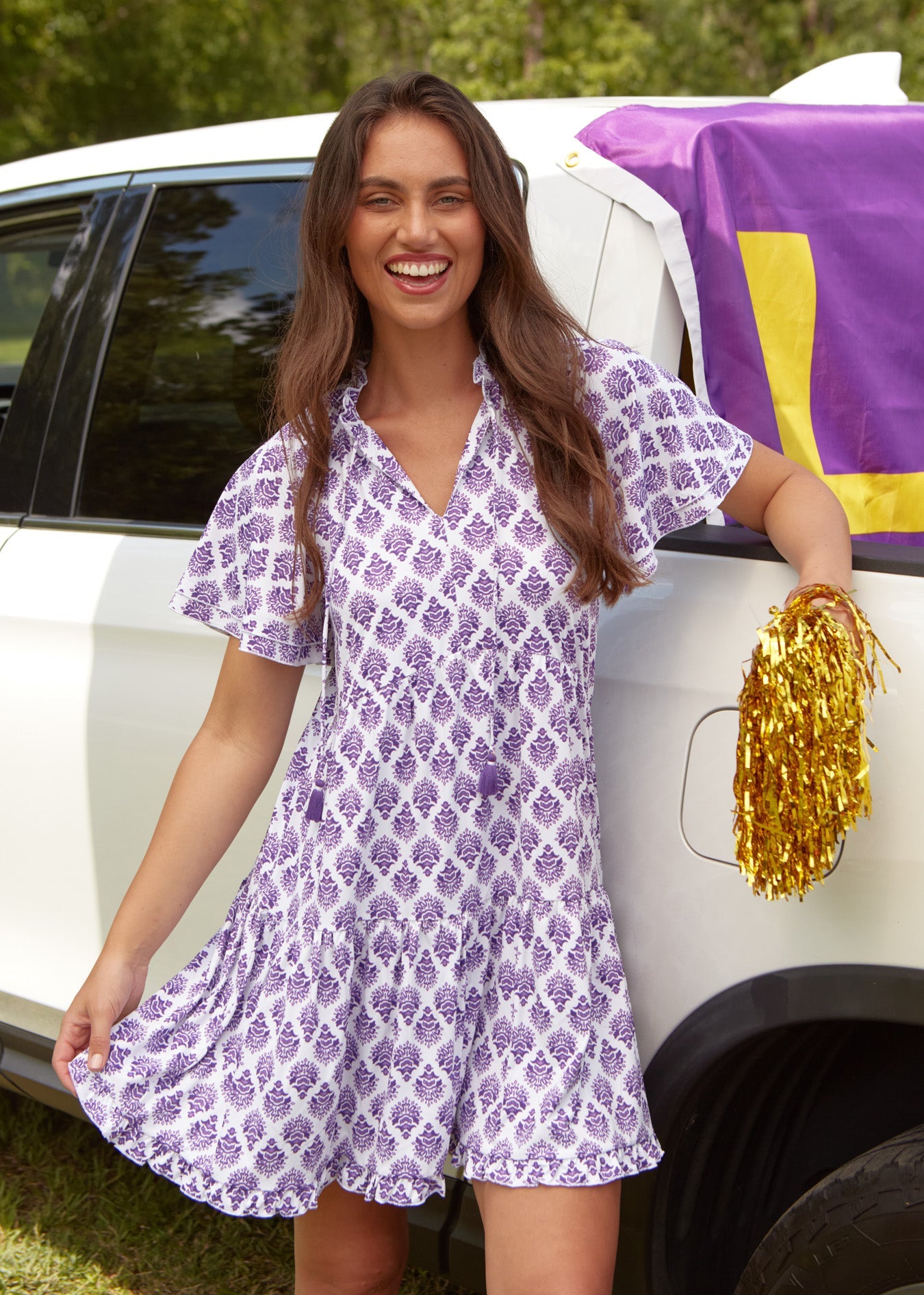 A brunette woman smiling & holding a gold pom pom while wearing the sun protective Cabana Life Purple Short Sleeve Tiered Dress in front of a white truck.