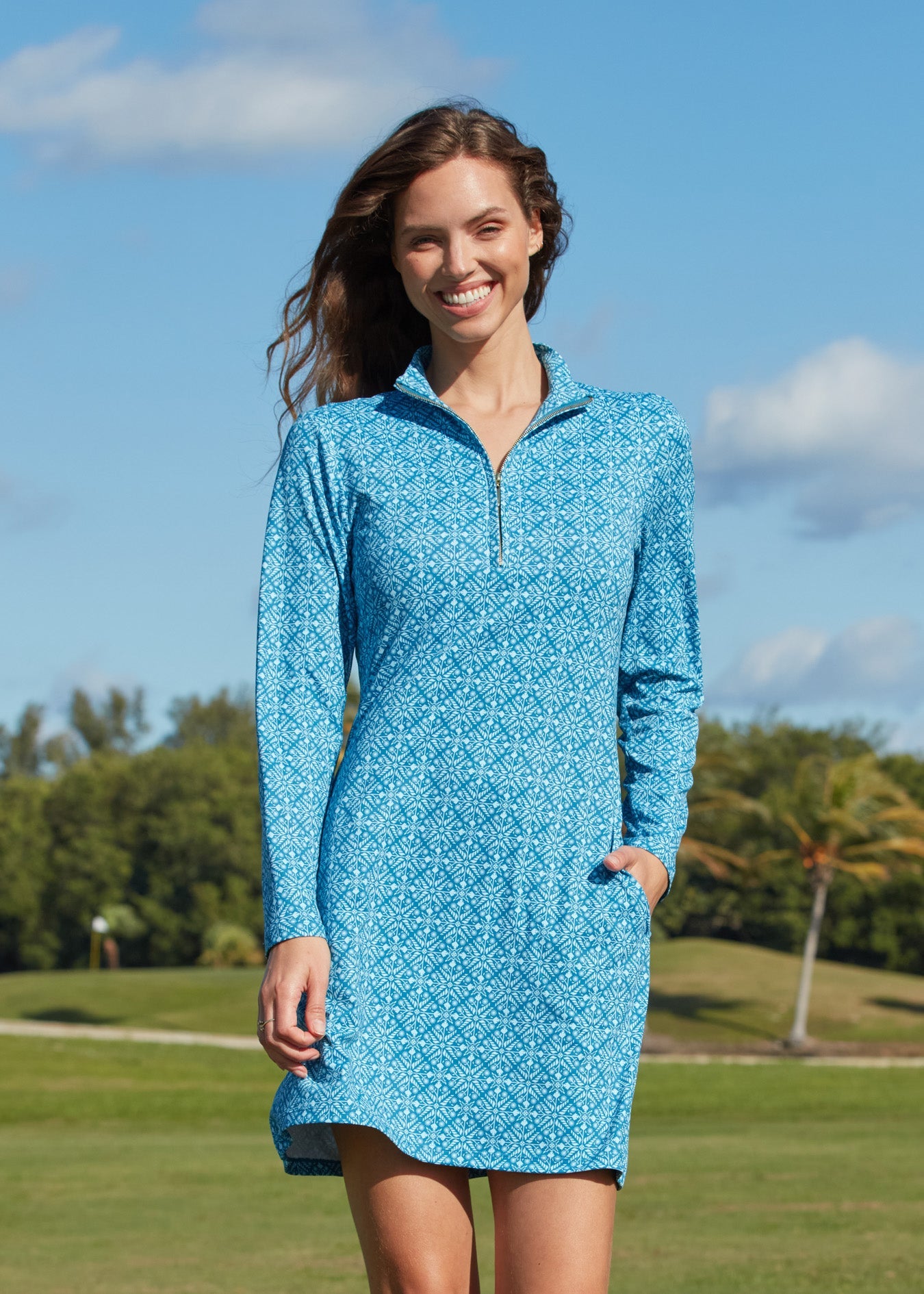 A brunette woman walking on a golf course with hand in pocket wearing the sun protective and turquoise printed Camden 1/4 Zip Sport Dress from Cabana Life.