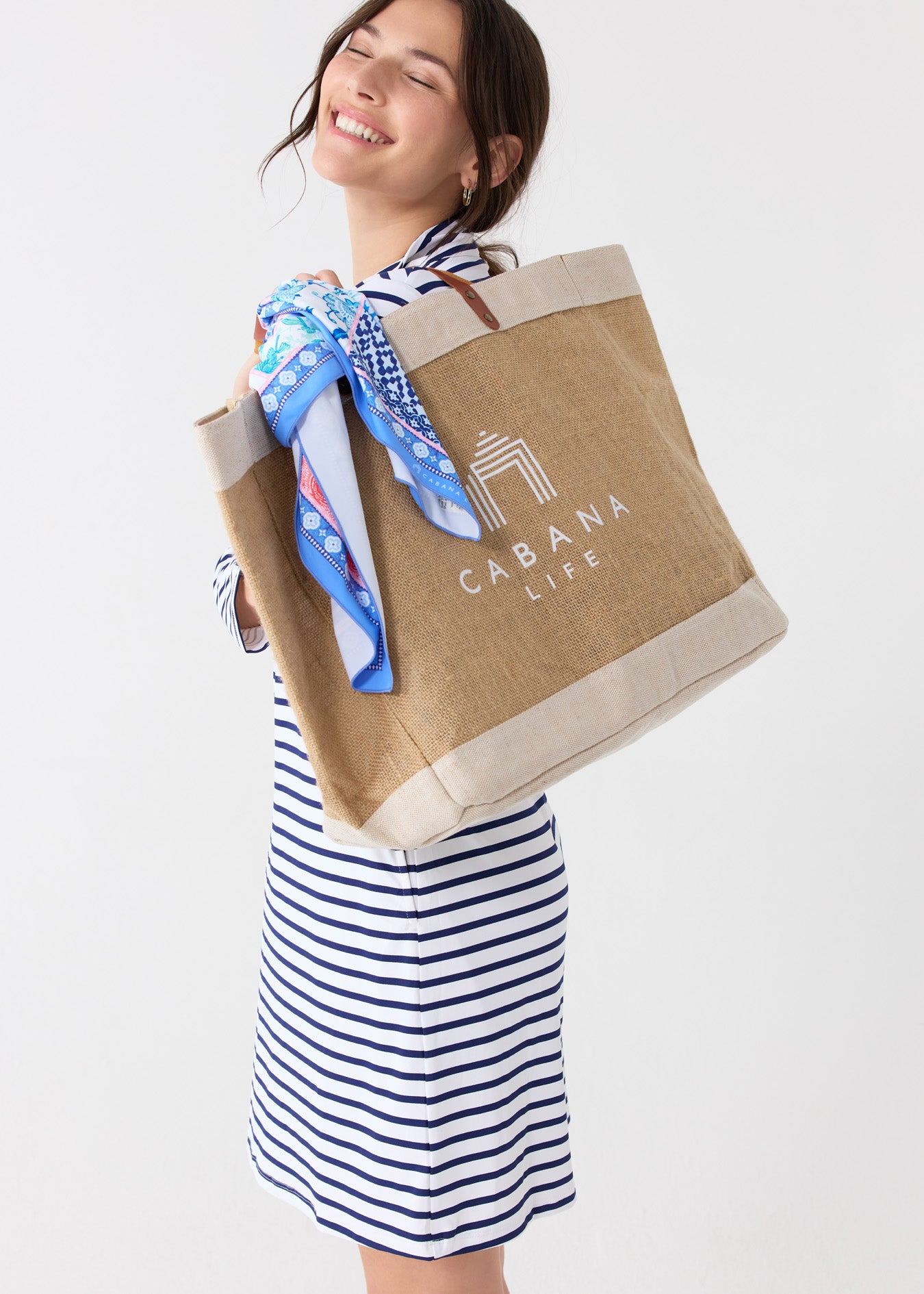 A woman wearing the Cabana Life sun protective Navy Stripe 1/4 Zip Sport Dress with the White Cabana Life Jute Bag over her shoulder tied with the 2025 Printed Patchwork Scarf.