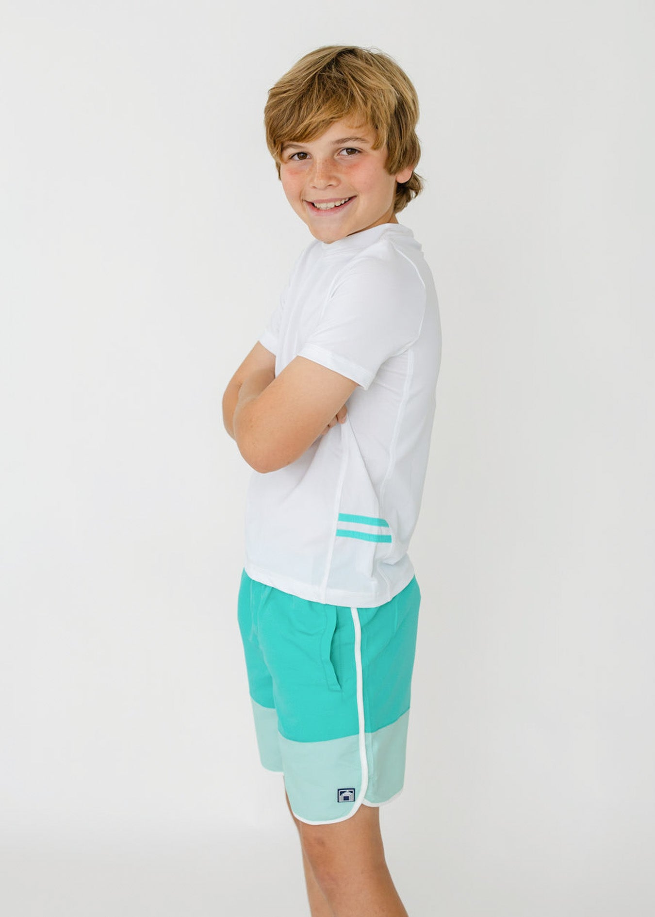 The side of a boy with his arms crossed wearing the sun protective Boys White Side Stripe Rashguard and Boys Aqua Colorblock Swim Trunk from Cabana life.