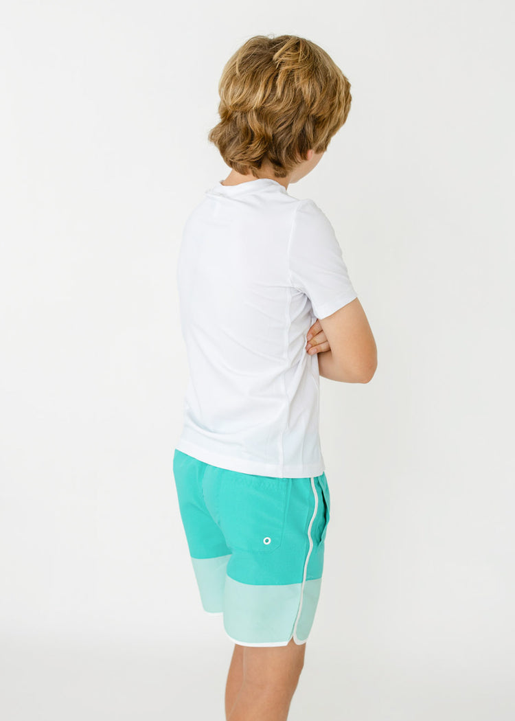 The back of a boy wearing the sun protective Boys White Side Stripe Rashguard and Boys Aqua Colorblock Swim Trunk from Cabana life.