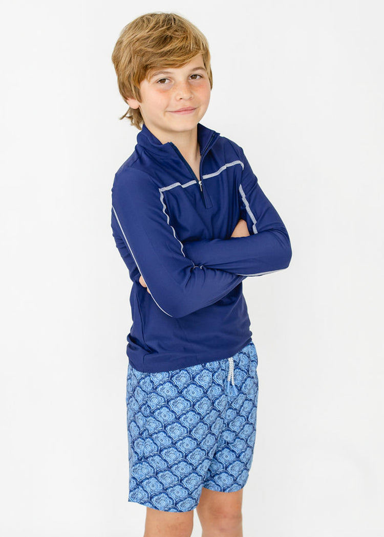 A boy wearing the sun protective Boys Navy Sport Zip Top and Boys Seaside Swim Trunk from Cabana Life with his arms crossed.