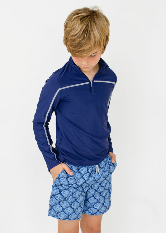 A boy wearing the sun protective Boys Navy Sport Zip Top with hands in pockets of the Boys Seaside Swim Trunk from Cabana Life.