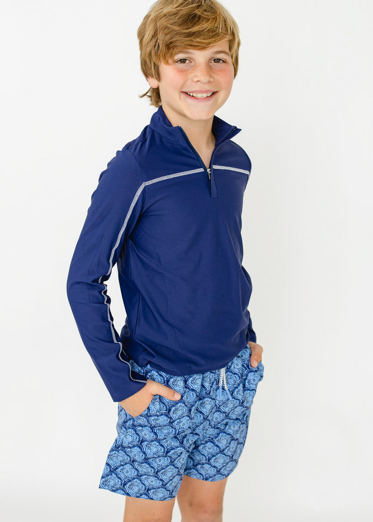 A boy wearing the sun protective Boys Navy Sport Zip Top with hands in pockets of the Boys Seaside Swim Trunk from Cabana Life.