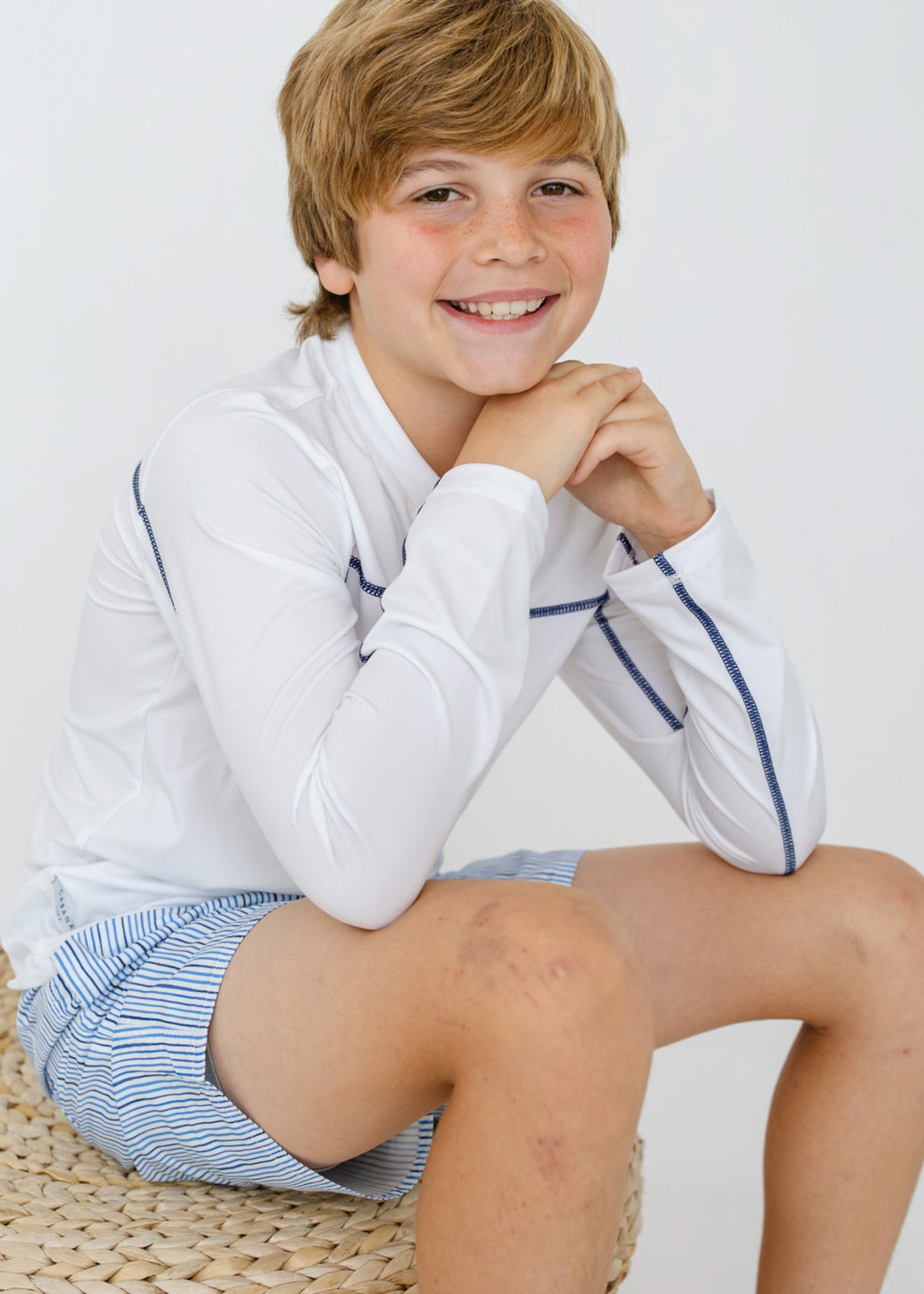 A boy sitting down wearing the sun safe Boys White Sport Zip Top and Boys Seaside Stripe Swim Trunk from Cabana Life.